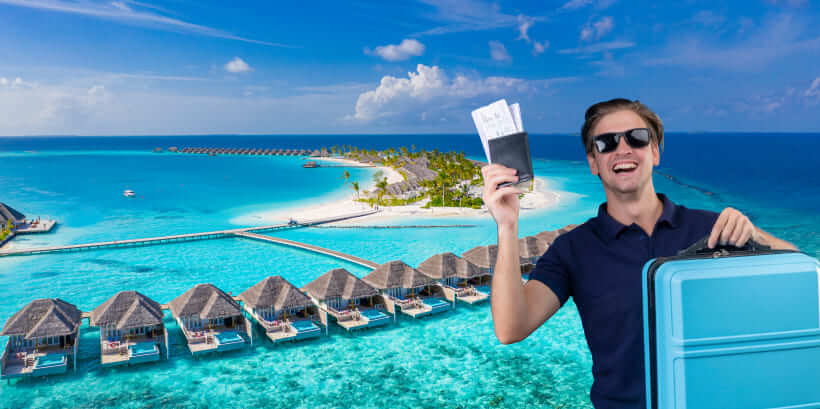 A smiling person in sunglasses holds a passport and boarding pass with one hand and a blue suitcase with the other. They stand against a backdrop of overwater bungalows and clear turquoise ocean in a tropical setting.