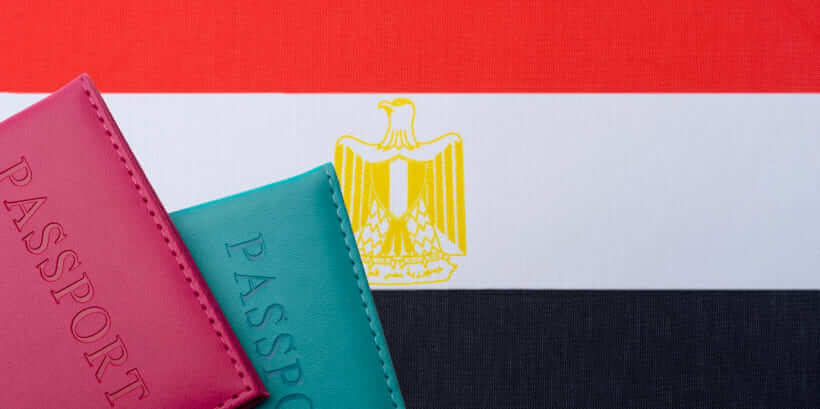 Two passports, one pink and one teal, partially visible in the foreground against the backdrop of the Egyptian flag with its red, white, and black stripes and a golden eagle emblem—a snapshot that hints at international adventures and perhaps a journey on how to apply for an Egypt visa from Dubai.