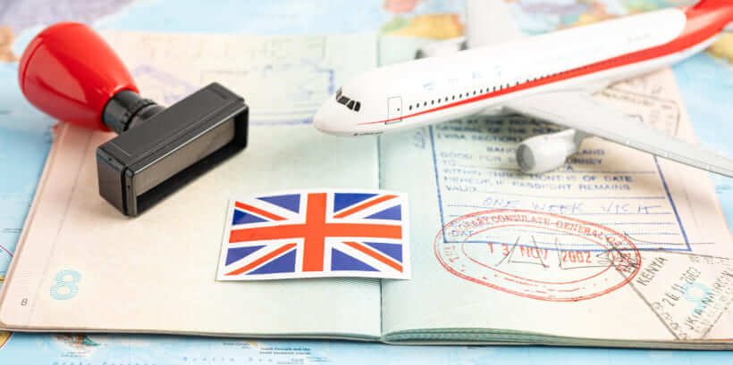 A passport with a UK multiple entry visa from Dubai, adorned with visa stamps, lies alongside a small airplane model and a British flag sticker on a world map, completed by the impression of a rubber stamp.